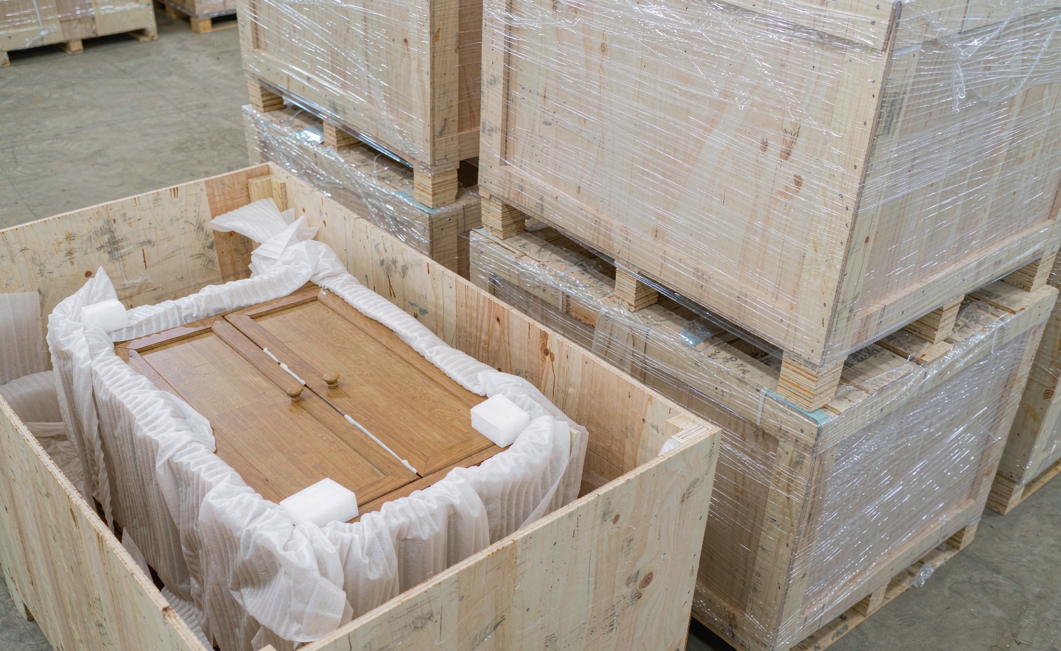 Armario envuelto en espuma de polietileno con esquineros blancos junto a cajas de madera contrachapada paletizadas en el almacén.